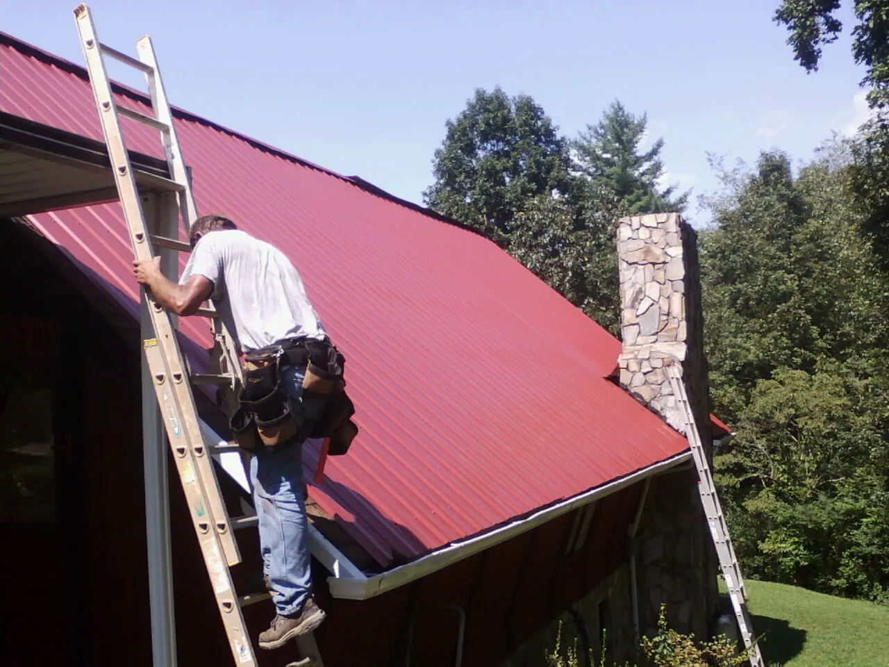 Roofer on a ladder beside a red metal roof and stone chimney — Soffit & Fascia Repair by Roofing Brothers in Welaka